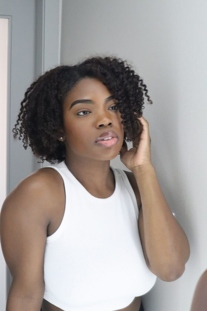 Woman with natural curly hair and glossy makeup looking at her reflection in a wall mirror.